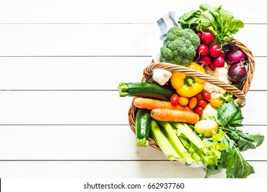 Colorful Organic Spring Vegetables In Wicker Basket - Captured From Above (top View). White Rustic Wooden Table As Background. Layout With Free Text (copy) Space. Fresh Harvest From The Garden.