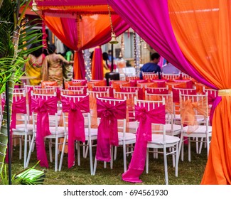 Colorful outdoor lawn tent and garden setting for Indian pre-wedding ceremony.  Vibrant pink and orange silk fabric curtains and bows on white chairs. Guests in background.