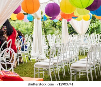 Colorful outdoor lawn tent and garden setting for Indian pre-wedding ceremony.  White silk fabric curtains. White chairs. Colorful round lanterns. Guests seated. Ivy decorations.