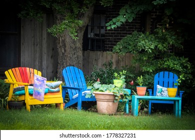 Colorful outdoor seating area - painted Adirondack chairs with pots of plants under tree in shady area and Home Sweet Home flag