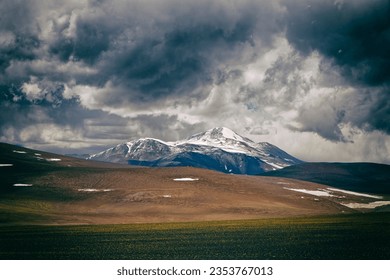 colorful panorama of snowy mountains overcast with dark blue clouds   with yellow and brown tones in the foreground. Cerro Bonete, Puna region, La Rioja, Argentina