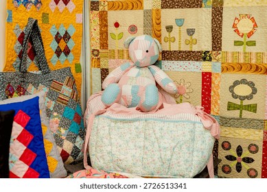 Colorful patchwork collection featuring a handmade teddy bear, a quilted tote bag, and pillows against a floral quilted background. Cozy nursery or home decor textile art