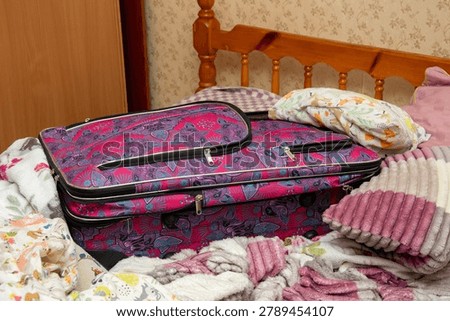 A colorful patterned suitcase sits packed on a cozy bed among pillows and soft blankets, suggesting packing, travel plans, and a relaxed home departure scene for vacation or short trip.