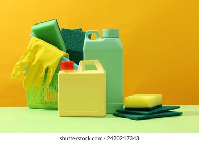 colorful photo for advertising cleansing products. Plastic canisters decorated with green plastic basket filled with cleaning supplies: rubber glove, sponges and brush on yellow background - Powered by Shutterstock