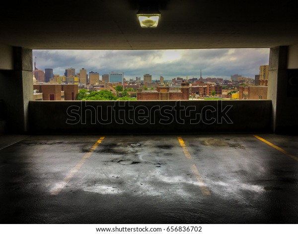 Colorful Photo of a View of the City of\
Baltimore from a Parking Garage - with a Wet, Rain Slick Floor and\
a Cloudy Blue Sky Over the Skyline in the\
Background