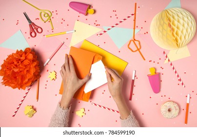 Colorful pink background with party confetti, paper decoration, flags, stationary with woman's hands putting greeting card in envelope. Flat lay top view. Making wish, sending letter invitation