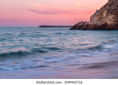 Colorful pink dreamy sunset over Atlantic Ocean coast on the beach of Albufeira city, Faro, Algarve, Portugal with soft long exposure water with limestone cliffs and lighthouse in the background