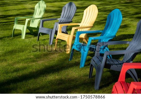Colorful plastic Adirondack chairs lined up on the grass of a yard in sunlight and shadows on a summer day.