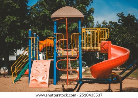 A colorful playground set with a slide, climbing wall, and a covered platform. The playground is surrounded by green trees and blue sky.