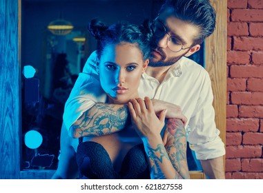 Colorful portrait of beautiful couple: brutal man in elegant suit and sexy girl with a tattoo wearing lingerie in barbershop
