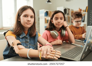 Retrato colorido de los niños que utilizan las computadoras en la Biblioteca de la escuela y mirando el foco de la cámara en la muchacha joven con la prótesis del brazo