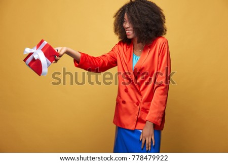 Colorful portrait of young african girl with afro hairstyle. Smiling girl wearing orange jacket, dark blue latex skirt holds red present in her hand and posing on yellow background. Studio shot.