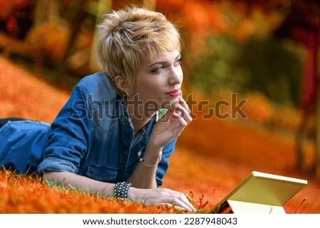Colorful portrait of a young woman lying face-down on the grass while using a laptop-tablet combo. In the midst of nature, well-off, she can afford to reflect on how long this will last before the cli