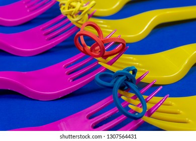 Colorful purple and yellow plastic forks with interlocking prongs and colored twisted rubber bands on top, arranged in a diagonal line on blue. Color trends and plastic waste problem concept - Shutterstock ID 1307564431