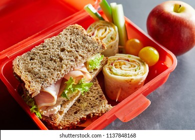 Colorful Red Plastic Kids Lunch Box Filled With Healthy Food Including Fresh Vegetables, A Wholewheat Ham Sandwich And Rolex Roll On Chappati With A Fresh Apple Alongside