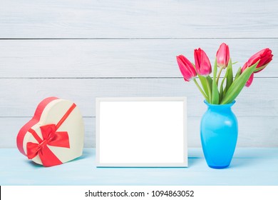 Colorful Red Spring Tulip Flowers In Nice Blue Vase And Blank Photo Frame With Decorative Heart Giftbox On Light Wooden Background As Greeting Card. Mothersday Or Spring Concept