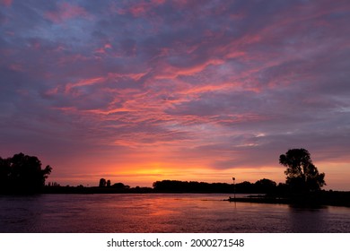 Colorful reflection in river IJssel with silhouetted trees of dramatic vibrant intense colorful sunset sky with texture and detail of cloud cover lighting up orange, red and magenta tones.