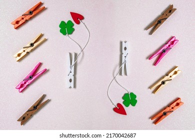 Colorful set of different wooden and plastic clothes pegs on a light pink glitter background. A red and a green lucky clover leaf are attached to each of the white wooden clothes pegs. Top view.