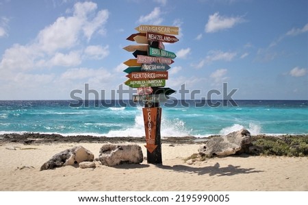 A colorful sign post with different locations and their distances from that point listed stands on a beach in Cozumel, Mexico with ocean waves crashing in the background under a clear blue sunny sky