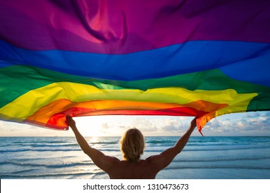 Colorful Silhouette Of A Man With Blond Hair Holding A Gay Pride Rainbow Flag Blowing In The Wind On A Tropical Beach With Golden Sun