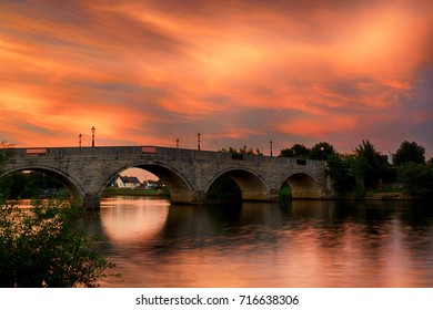 Colorful sky at sunset over Chertsey Bridge, Surrey, UK. Beautiful red, yellow, orange and pink clouds reflected in the River Thames as it flows under the stone built arches of the old bridge.