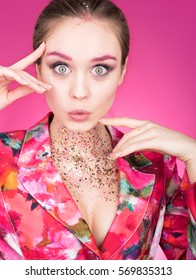 Colorful Studio portrait of a beautiful young woman with eyes wide open. Much surprised look. Makeup with glitter. Depict and gesticulating hands. Pink background