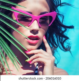 Colorful summer portrait of young attractive brunette woman wearing sunglasses under a palm tree by the swimming pool 