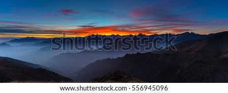 Colorful sunlight behind majestic mountain peaks of the Italian - French Alps, viewed from distant. Fog and mist covering the valleys below, autumnal landscape, cold feeling.
