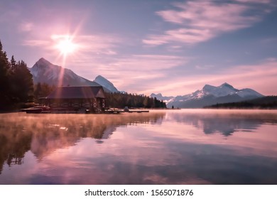 Colorful sunrise on famous Maligne lake with boat house and white snowy mountains in background. Moody, soft, pink colors of the morning sky, mirror reflection on the lake. Jasper National Park Canada