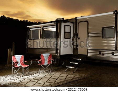 Colorful sunset behind a brightly lit camping trailer with chairs outside