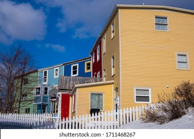 Colorful tall buildings of yellow, red, blue and green colour. There are fences and snow in the small yards with shrubs and trees under blue sky. The houses have double hung windows on each floor.  