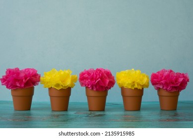 Colorful Tissue Paper Flowers In Pink And Yellow Are Captured In Small Terra Cotta Pots On A Weathered Table.