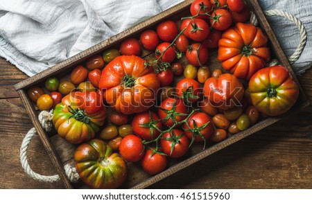 Colorful tomatoes of different sizes and kinds in dark wooden tray over light textile and rustic wooden background, top view, horizontal composition