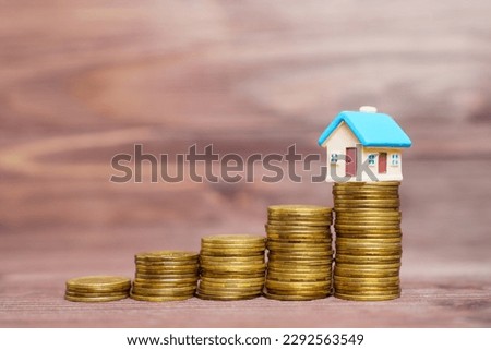 Colorful toy house figure standing on top of a pile of coins, all set against a wooden background. Personal finance, investing, real estate and wealth management related concept.