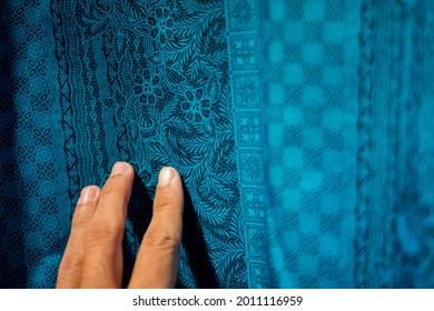 A colorful traditional Indonesian fabric named "batik" with floral motif on black ink on a blue background, held by a man's left hand