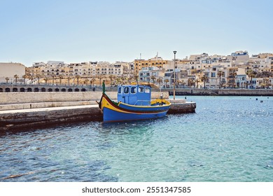 Colorful traditional Maltese fishing boat docked in calm harbor with backdrop of Mediterranean-style buildings under clear blue sky - Powered by Shutterstock