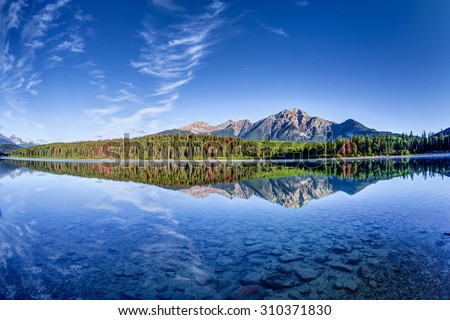 Colorful trees lined the shores of Patricia Lake at Jasper National Park with Pyramid Mountain in the background. The calm lake reflects a mirror image of the mountains and trees.