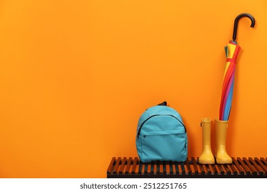 Colorful umbrella, backpack and rubber boots on wooden bench near orange wall, space for text - Powered by Shutterstock