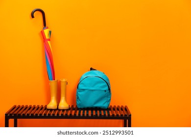 Colorful umbrella, backpack and rubber boots on wooden bench near orange wall, space for text - Powered by Shutterstock