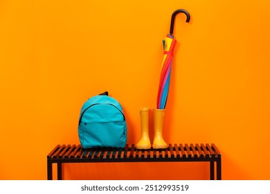 Colorful umbrella, backpack and rubber boots on wooden bench near orange wall - Powered by Shutterstock