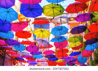 Colorful umbrellas suspended in the air above a narrow street. Umbrellas are in shades of blue, red, pink, purple, and yellow, creating a canopy effect. Artistic and engaging urban scene - Powered by Shutterstock