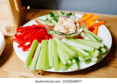 Plato de tentempiés de verduras coloridas con palos de apio frescos, palos de zanahoria, pimiento de campana roja y cremoso humus sumergido en un recipiente de vidrio.