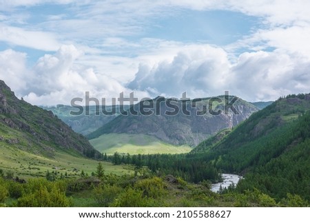 Colorful view to sunlit green mountain valley with forest and river against mountain range under cloudy sky. Wide mountain valley in sunlight and large mountains in lush clouds in changeable weather.