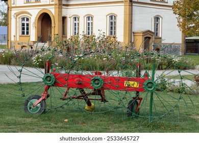 Colorful vintage agricultural rake with large green wheels displayed in front of an old manor house. Concept of farming equipment, rural heritage, and agricultural machinery in a historical setting - Powered by Shutterstock