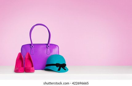 Colorful Woman Fashion Accessories On The Table Isolated On Pink. A Purple Leather Bag,red Heels And A Blue Green Hat.