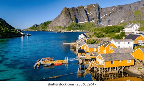 colorful wooden cabins perched on stilts over the clear blue waters of a Norwegian fjord, surrounded by lush green hills and majestic mountains. Sakrisoy, Lofoten, Norway - Powered by Shutterstock