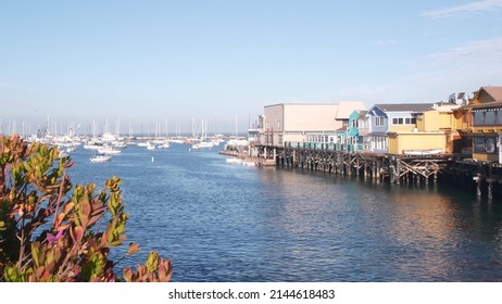 Colorful wooden houses on piles or pillars, ocean bay or harbor, sea water. Old Fisherman's Wharf. Yachts, sail boats in Monterey Marina, California coast USA. Tourist beachfront promenade, flowers.
