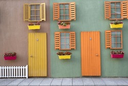 Colorful Wooden Yellow And Orange Door With Windows On Green And Brown Wall, White Fence And Flowers In Pots, Shutters Open. Decorative Vintage House Concept