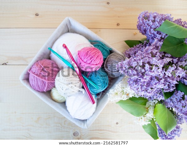 colorful woolen balls with\
crochet hook in a basket on wooden ground with purple lilac flower\
spring