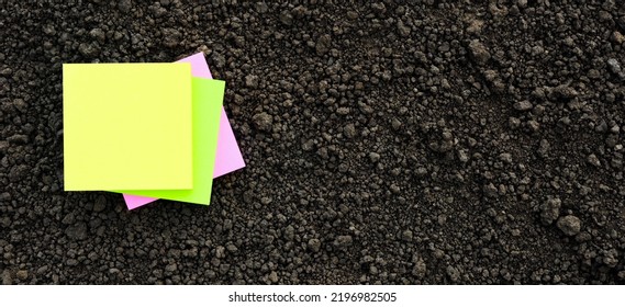 Colors Note Paper Square On Black Soil Texture Ground Close Up. Yellow Sticky Note Blank Paper Flat Soil Organic Earth Texture. Mockup On Ground Soil Background Top View From Above. Agriculture Banner